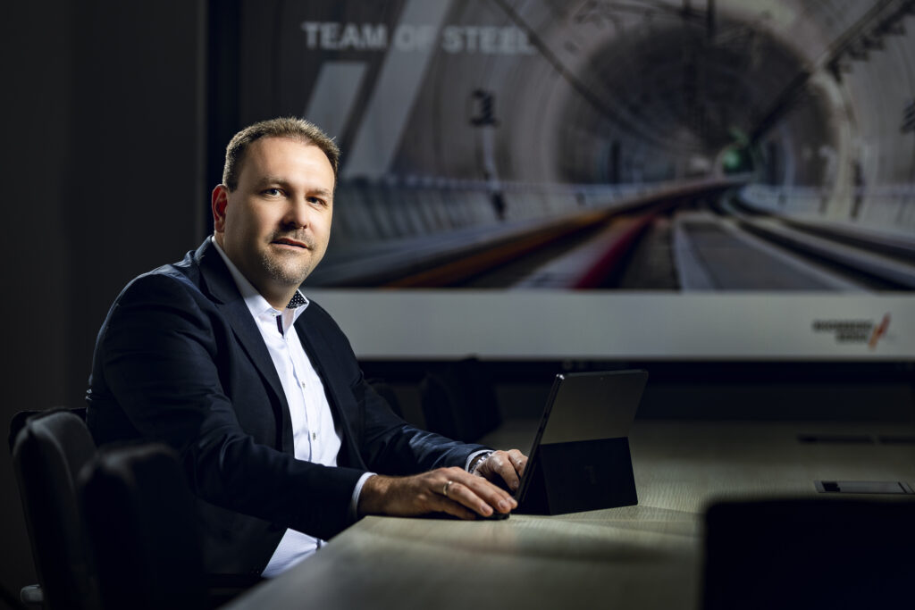 Ein Mitarbeiter im dunklen Anzug mit Krawatte vor einer großformatigen Fotografie.