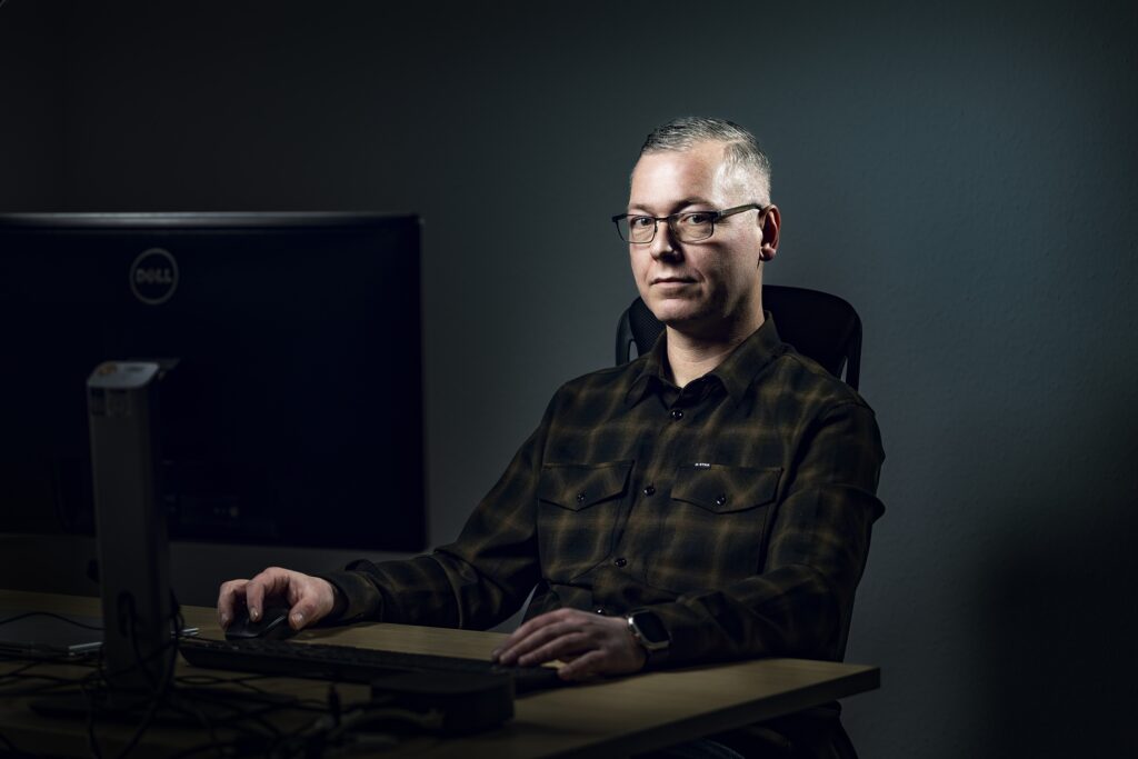 Ein Mitarbeiter mit Brille und dunklem Hemd vor einer grau beleuchteten Wand.