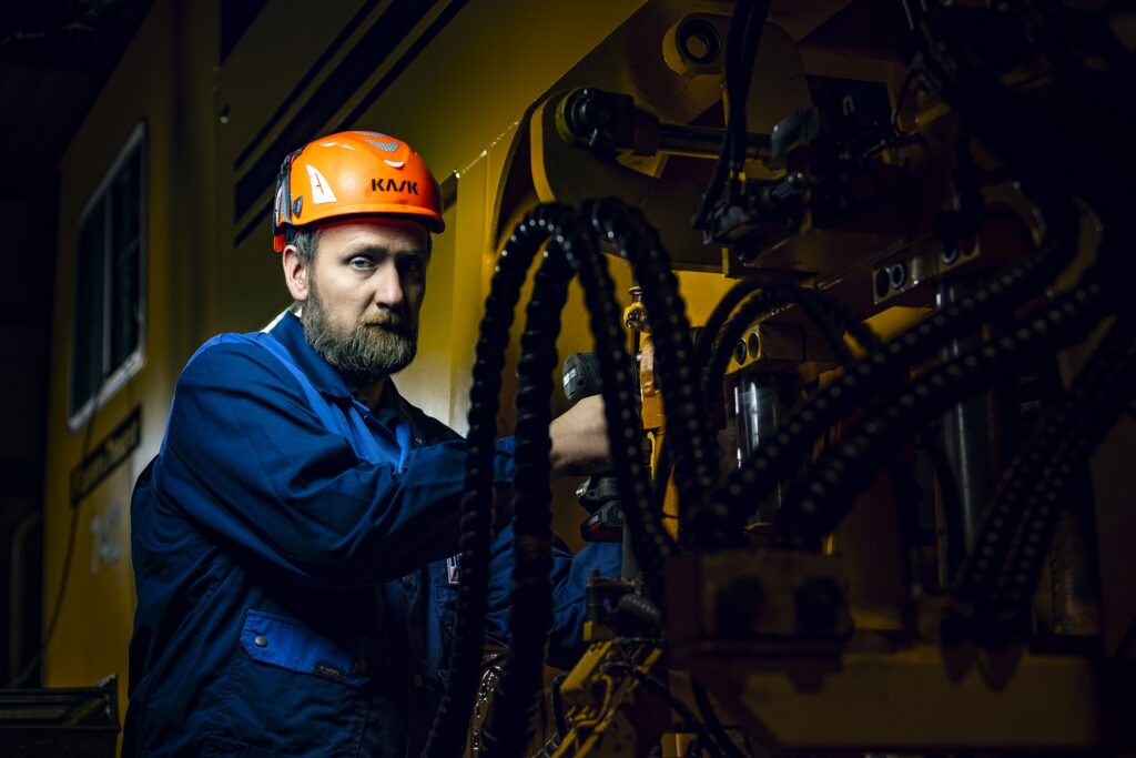 Ein Mitarbeiter in blauer Arbeitskleidung und orangefarbenem Schutzhelm neben einer gelben Maschine.