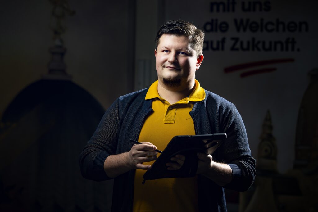 Ein lächelnder Mitarbeiter in gelbem Poloshirt und dunkler Jacke vor einer Wand mit Schrift.