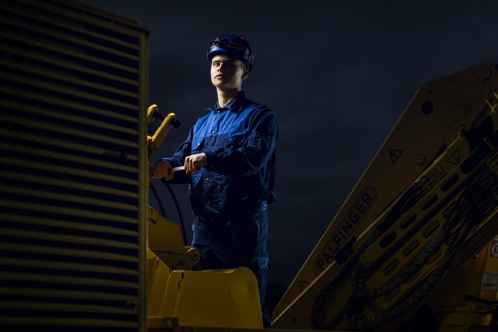 Ein Mitarbeiter in blauer Arbeitskleidung und dunkler Kopfbedeckung vor einem dunklen Werkstatthintergrund.
