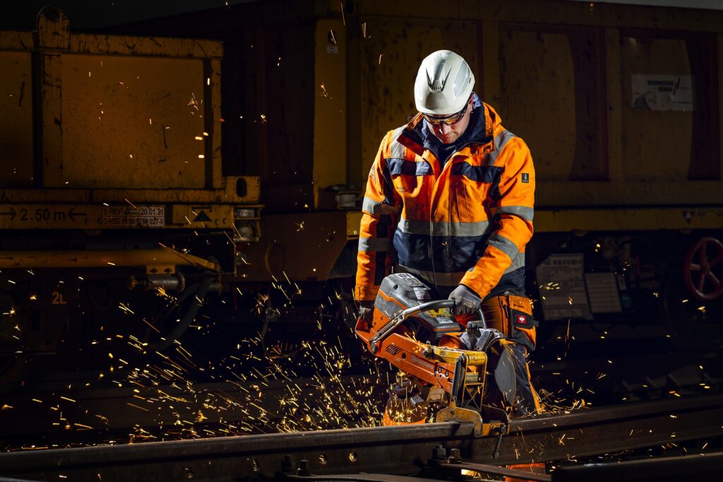 Ein Mitarbeiter mit weißem Schutzhelm beugt sich in orangefarbener Warnkleidung über seinen Arbeitsbereich.
