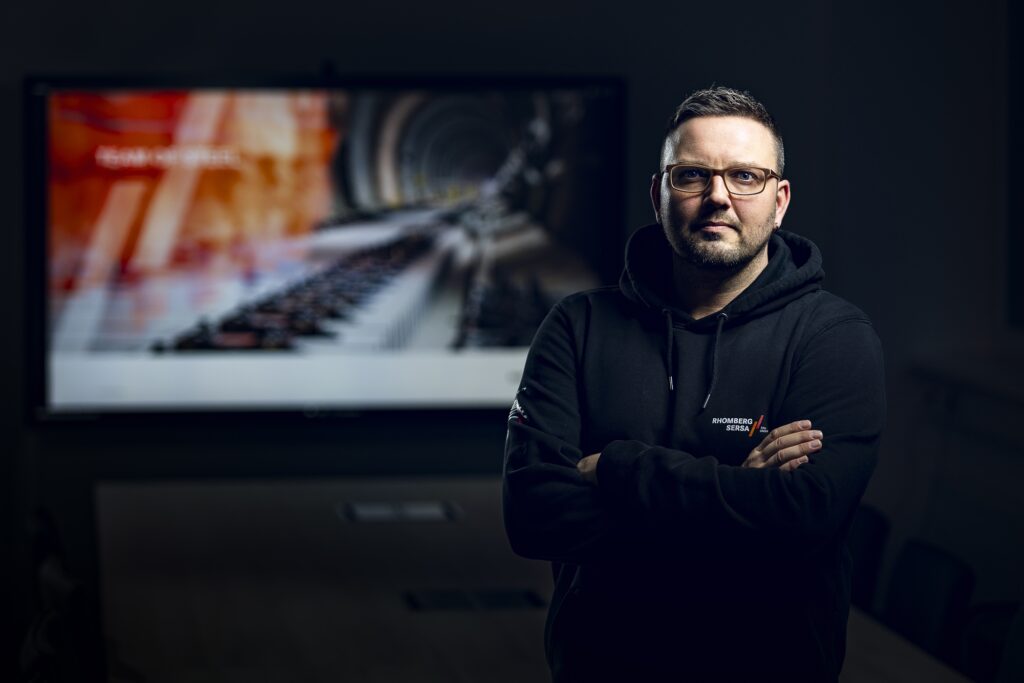 Ein Mitarbeiter mit Brille, Bart und dunklem Oberteil vor einer großformatigen Fotografie.