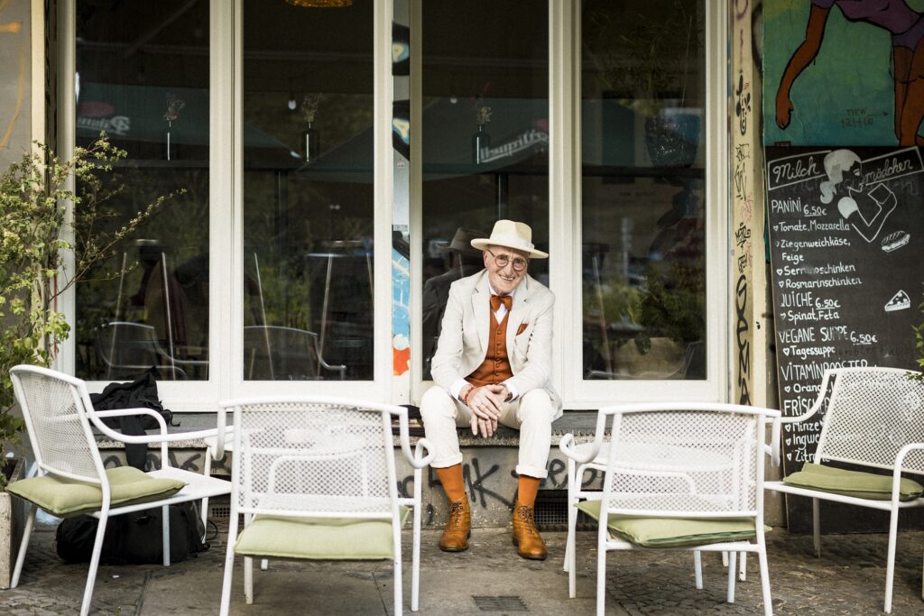 Günther Krabbenhöft sitzt lächelnd vor einem Café zwischen weißen Stühlen, mit hellem Hut und orangefarbenen Socken.