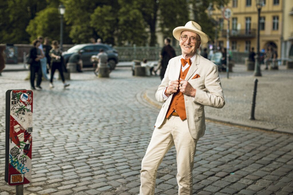 Günther Krabbenhöft steht lächelnd im hellen Anzug und Hut auf einer gepflasterten Berliner Straße und hält die Revers seines Sakkos.