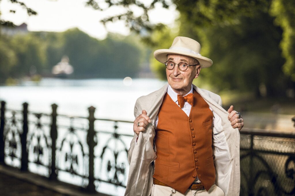 Günther Krabbenhöft öffnet sein helles Sakko und zeigt eine orangefarbene Weste mit passender Fliege, im Hintergrund das Wasser.