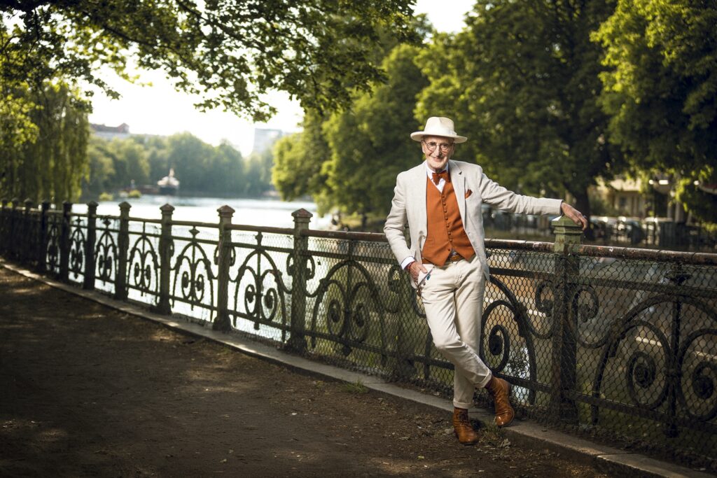 Günther Krabbenhöft lehnt mit gekreuzten Beinen an einem verzierten Geländer am Wasser, eingerahmt von grünen Bäumen.