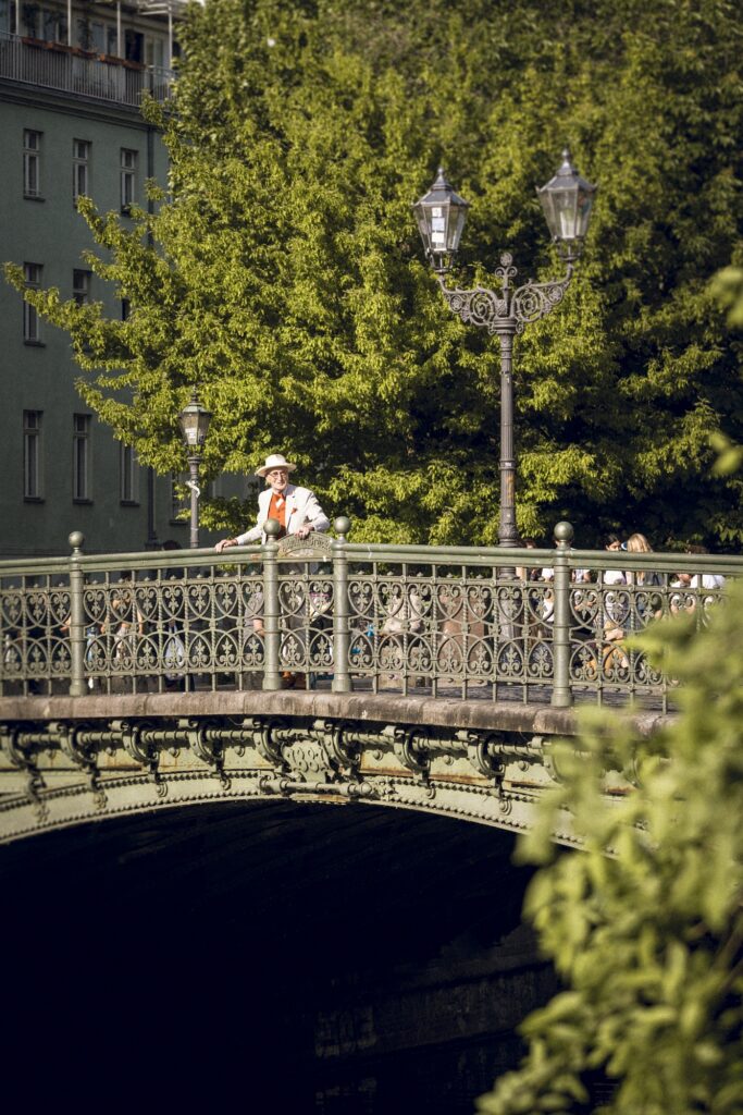 Günther Krabbenhöft steht auf einer verzierten Brücke unter historischen Laternen, umgeben von grünen Bäumen.