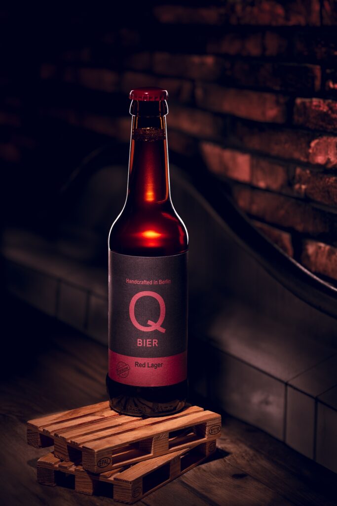 Eine braune Qbier-Flasche mit rotem Etikett vor einer dunklen Backsteinwand.