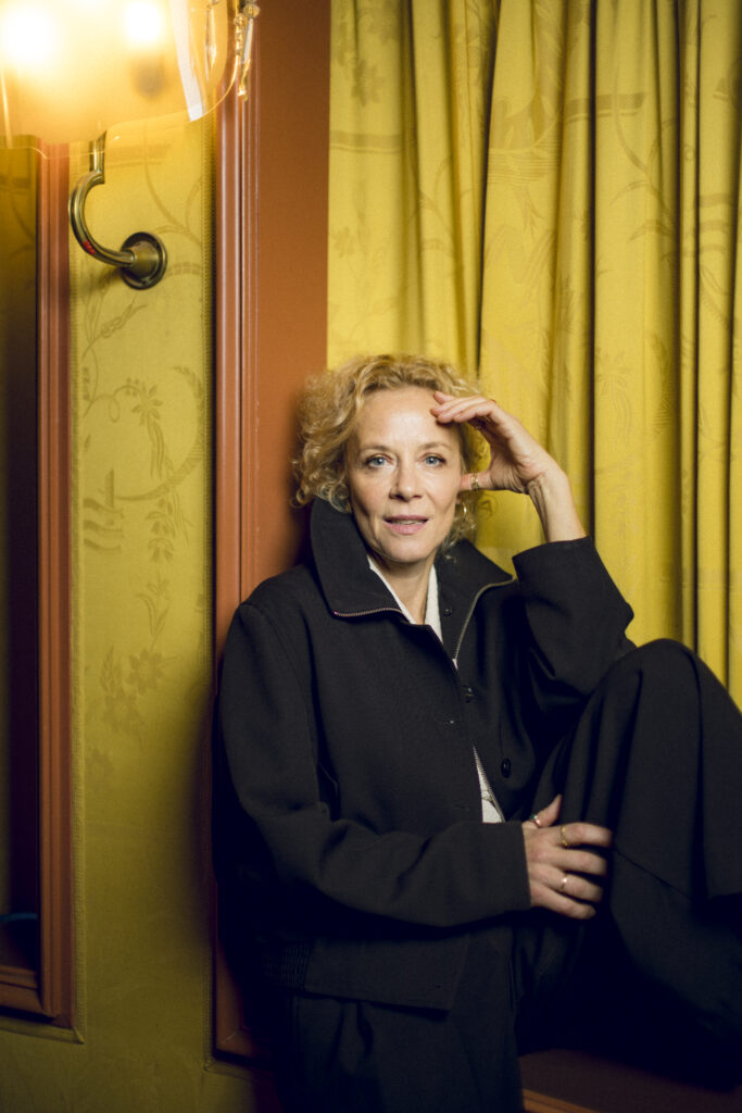 Katja Riemann in dunkler Kleidung vor einem gelben Vorhang, eine Hand an der Schläfe.
