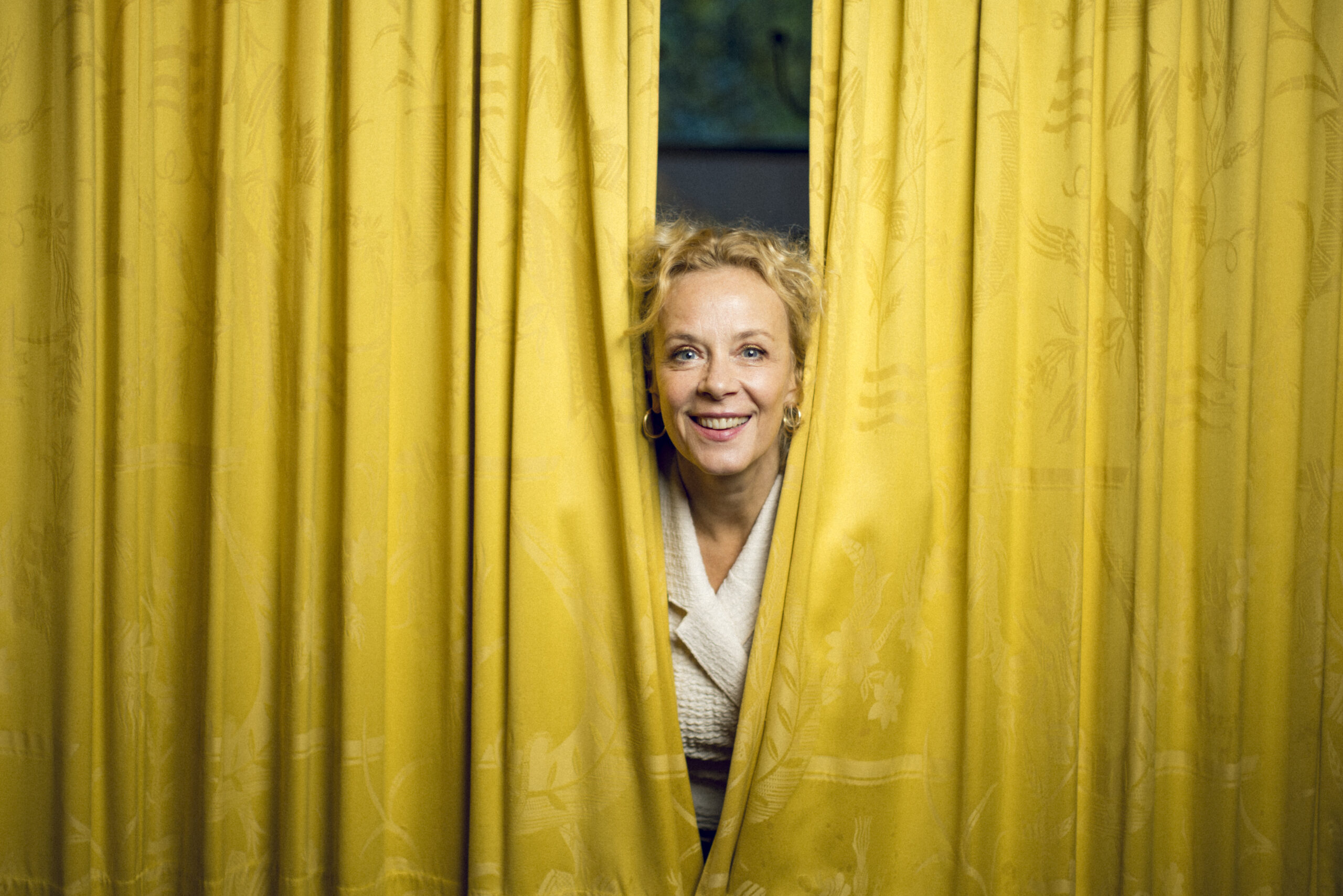 Katja Riemann blickt lächelnd zwischen zwei gelben Vorhängen hervor.