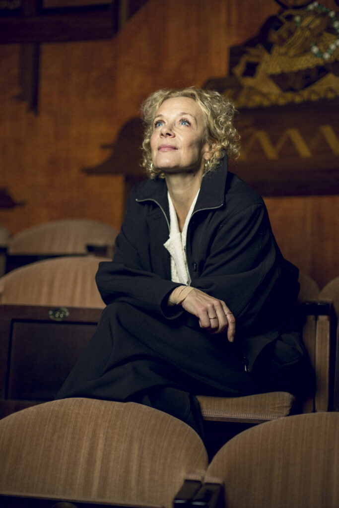 Katja Riemann sitzt in einem gepolsterten Sessel und blickt seitlich nach oben.