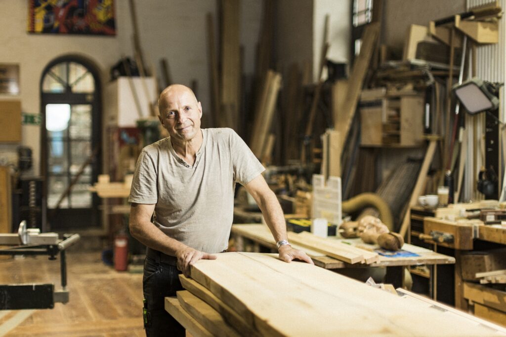 Ein Tischler steht hinter seiner Werkbank, umgeben von Holz und Werkzeugen.