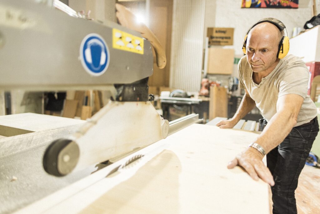 Ein Tischler arbeitet an einer hellen Holzfläche; im Vordergrund liegt unscharf weiteres Holz.