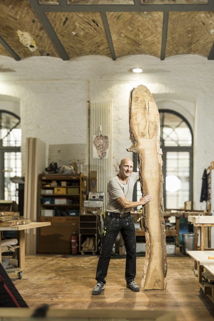 Ein Tischler steht mit einem hohen, naturkantigen Holzbrett in der Werkstatt.
