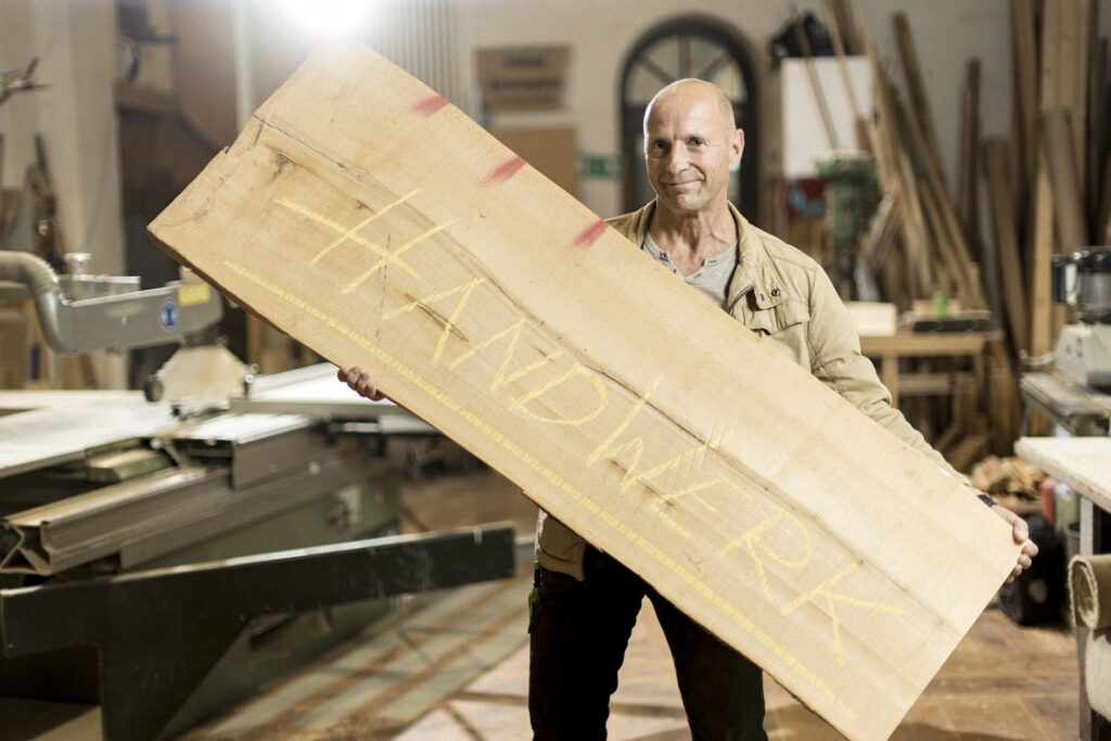 Ein Tischler hält eine große Holzplatte schräg vor sich in der Werkstatt.