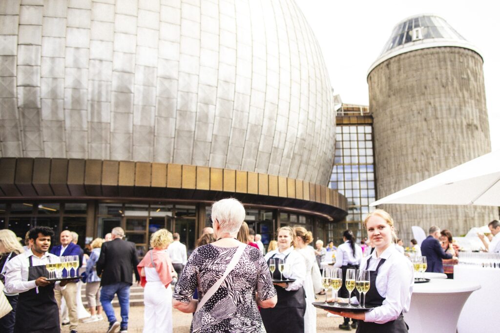 Gäste sitzen an Tischen vor einer großen gerundeten Metallfassade.