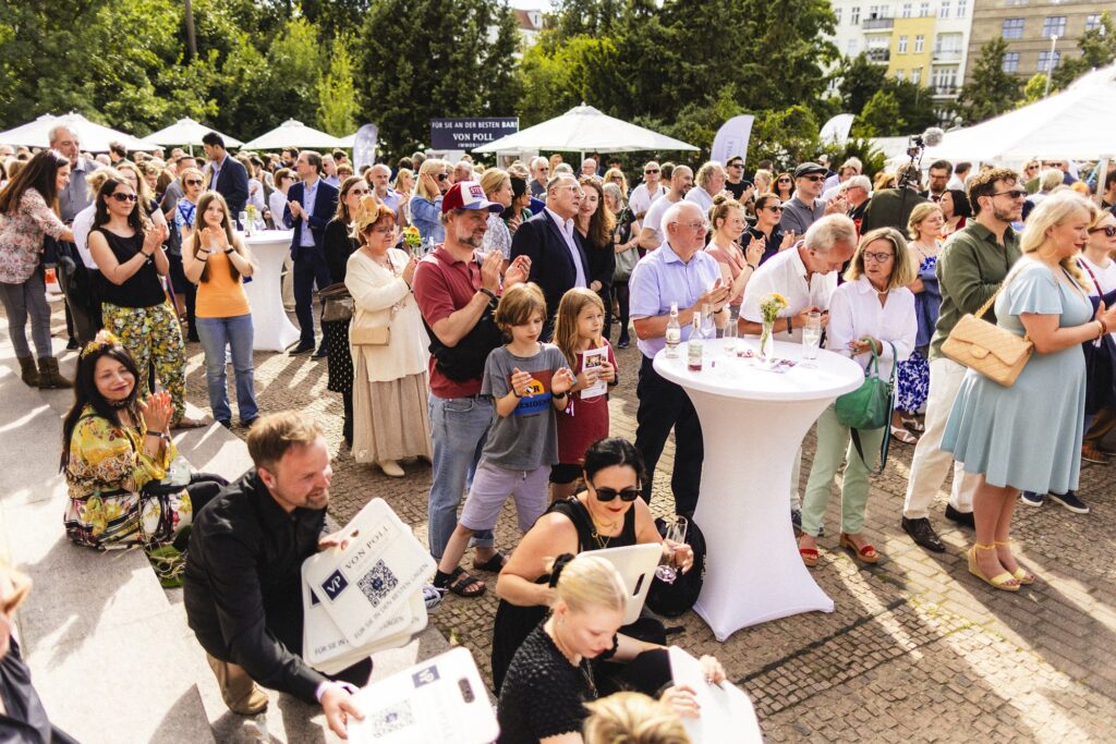 Besucher stehen und sitzen auf dem belebten Festgelände zwischen Bäumen und weißen Zelten.