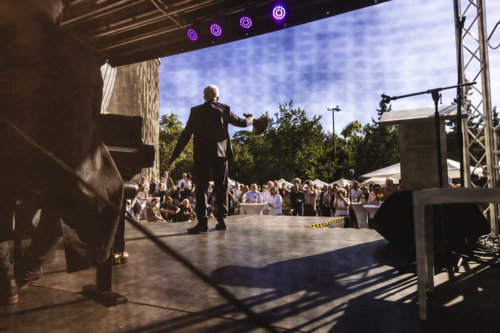 Eine Person steht auf einer überdachten Bühne vor dem Publikum des Sommerfests.