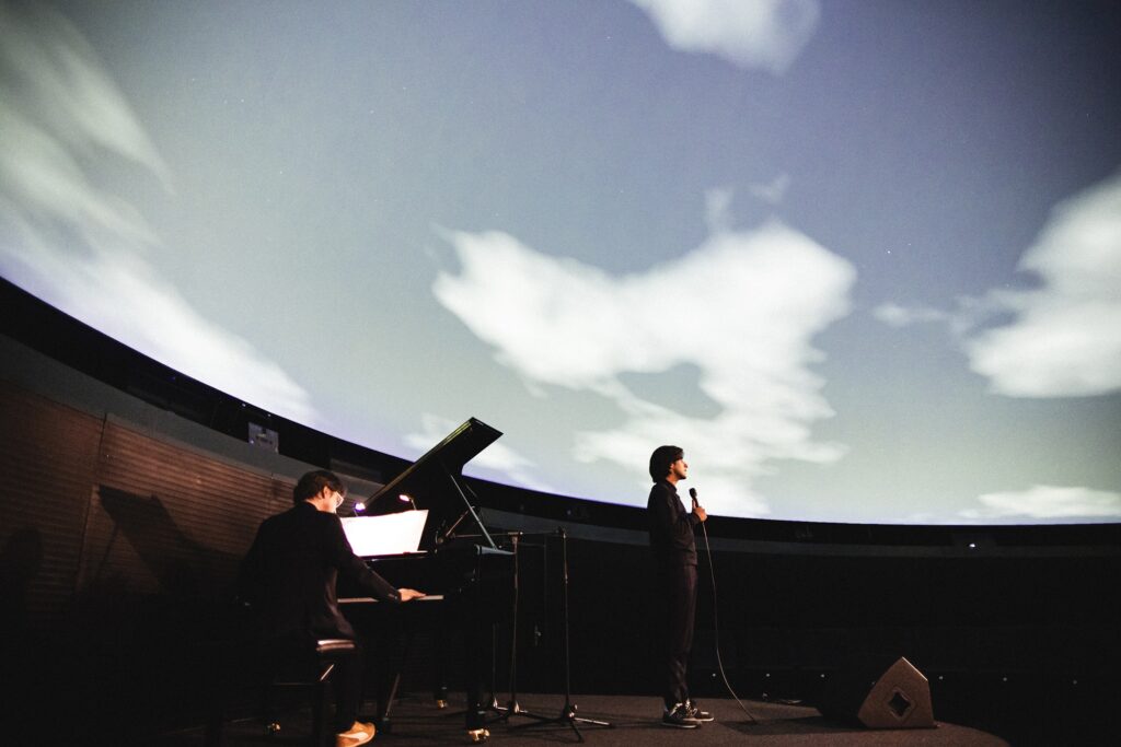 Eine große Leinwand zeigt Wolken am Himmel; davor sind Personen als Silhouetten zu sehen.