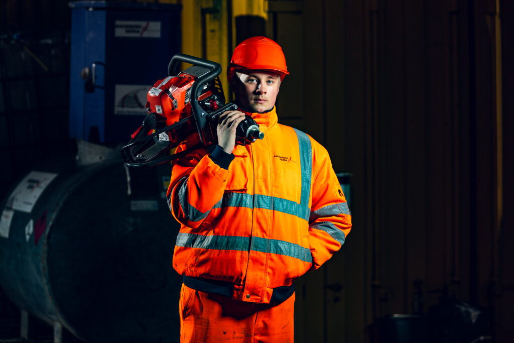 Ein Mitarbeiter mit orangefarbenem Schutzhelm trägt ein Werkzeug auf der Schulter.