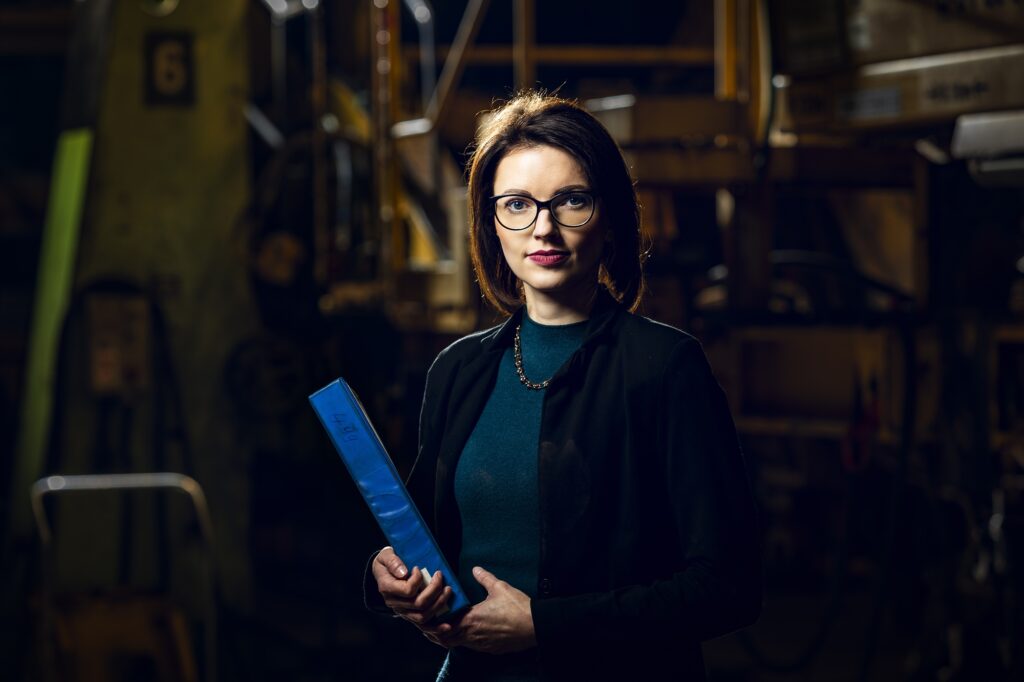 Eine Mitarbeiterin mit Brille und grünem Oberteil steht vor Maschinen in der Werkhalle.