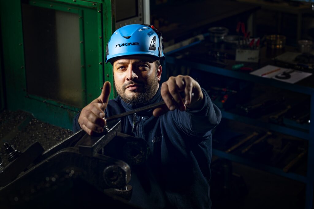 Ein Mitarbeiter mit blauem Schutzhelm hält seine Arbeitshandschuhe auf Schulterhöhe.