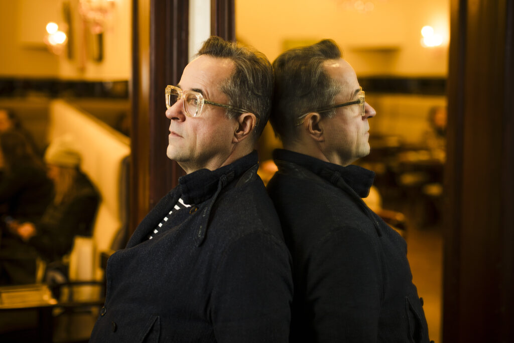 Jan Josef Liefers mit Brille im Profil, gespiegelt in einer Glasfläche.