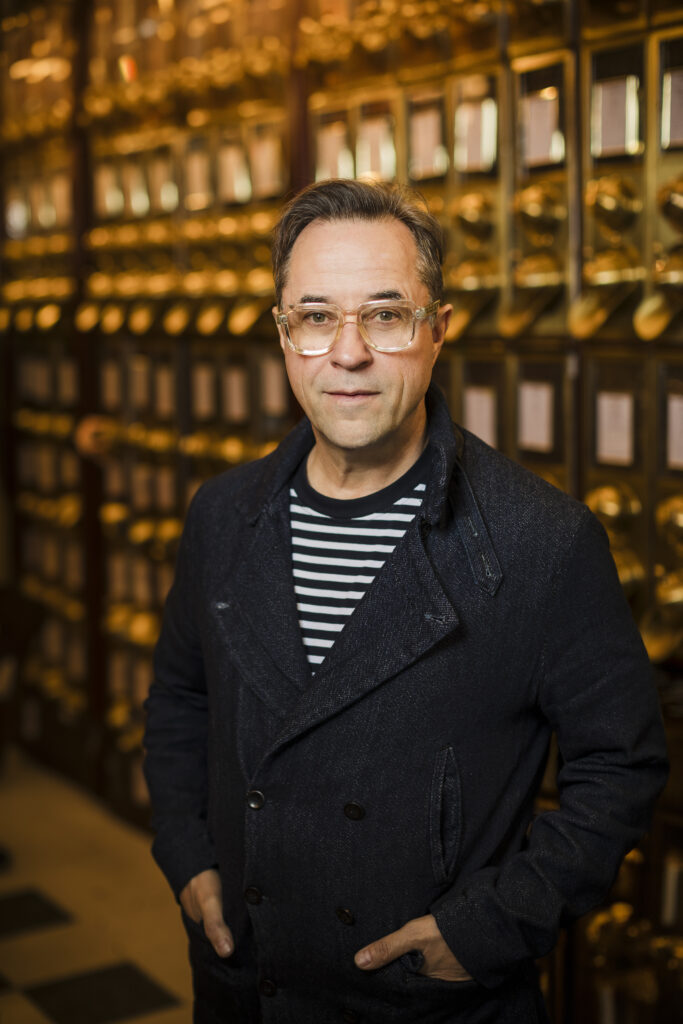 Jan Josef Liefers mit Brille, dunkler Jacke und gestreiftem Oberteil, frontal porträtiert.