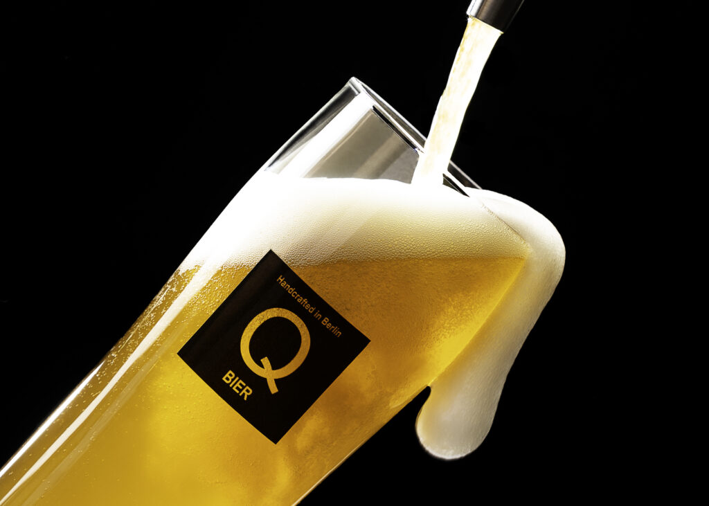 Helles Bier wird in ein schräg gehaltenes Glas mit Qbier-Logo eingeschenkt.