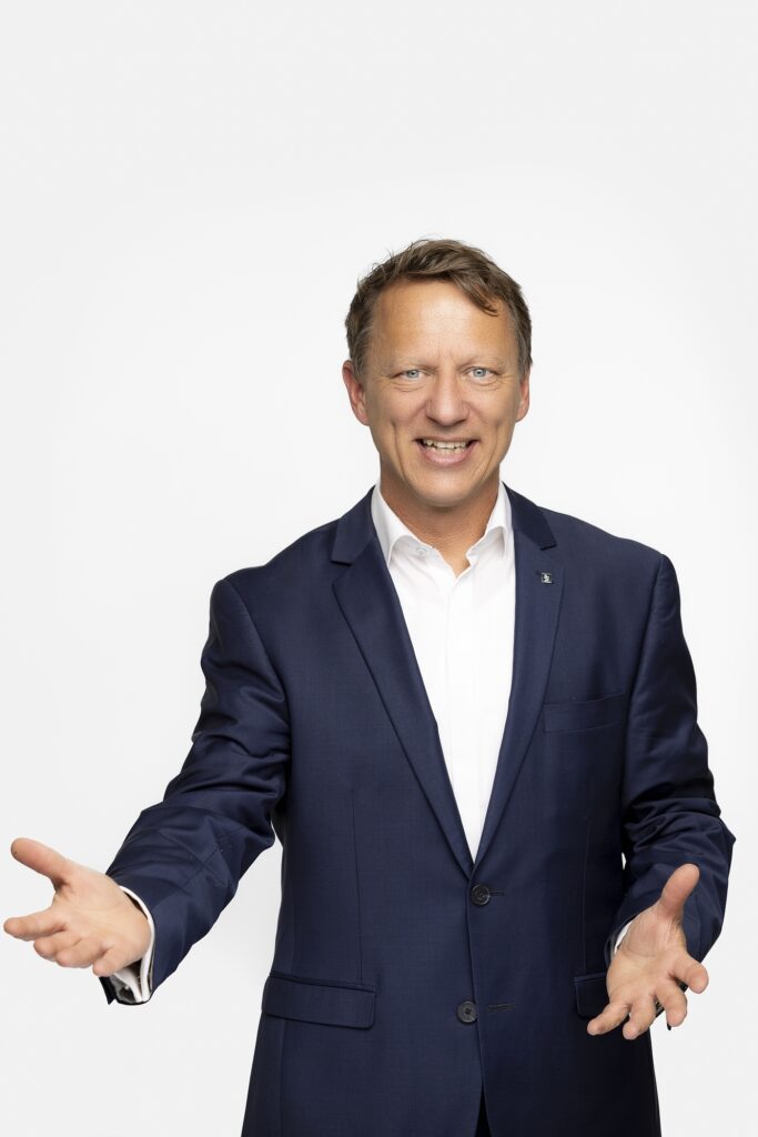 Ein lächelnder Mitarbeiter von VON POLL IMMOBILIEN im dunklen Sakko und weißen Hemd vor hellem Hintergrund.