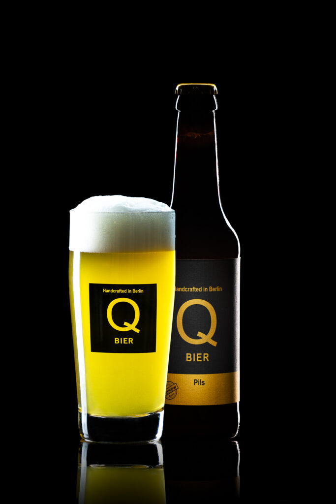 Ein gefülltes Bierglas mit Schaumkrone steht neben einer Qbier-Flasche vor schwarzem Hintergrund.